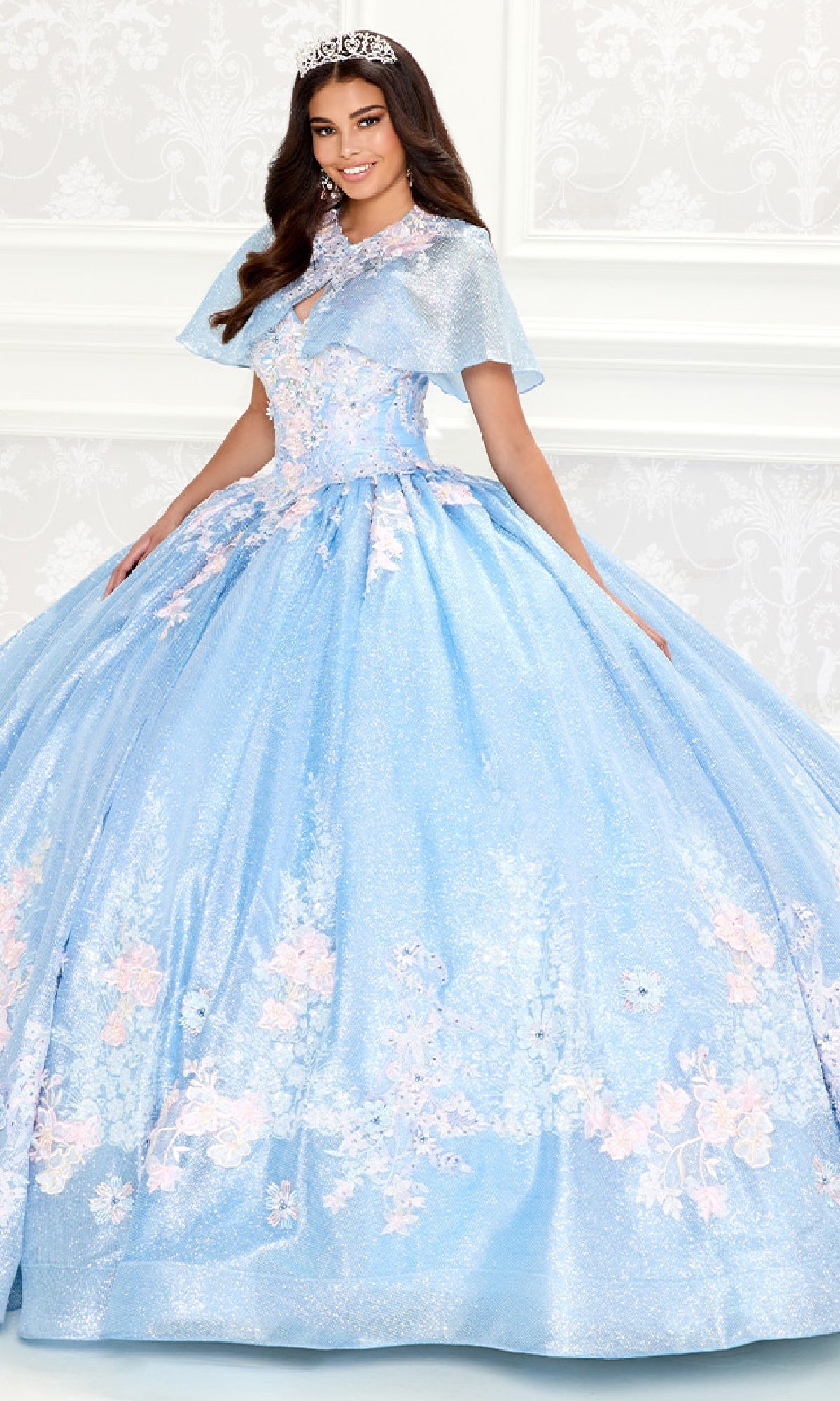 A young woman wearing the Princesa PR30083 Light Blue Quinceanera Dress with a corset bodice, floral details, and a tiara stands in front of a white patterned wall.